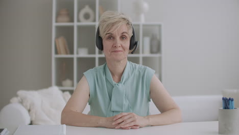 middle-aged-kind-and-friendly-woman-is-listening-by-headphones-and-nodding-sitting-at-table-and-looking-to-camera